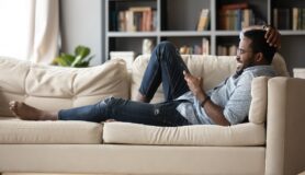 Full-length relaxed young man in glasses lying on comfortable couch, enjoying spending weekend leisure time chatting in social network on phone with friends