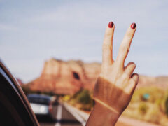 Woman holds two fingers out the window of a moving car while traveling as a digital noamd.