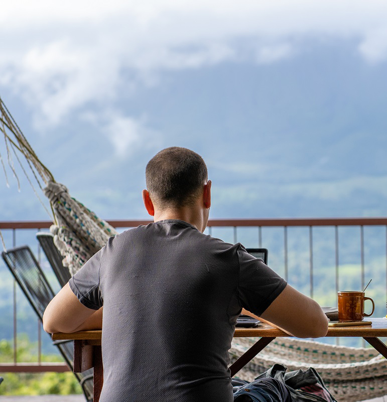 Digital nomad sitting on a table with breakfast while working with a view of a volcano in front of him.