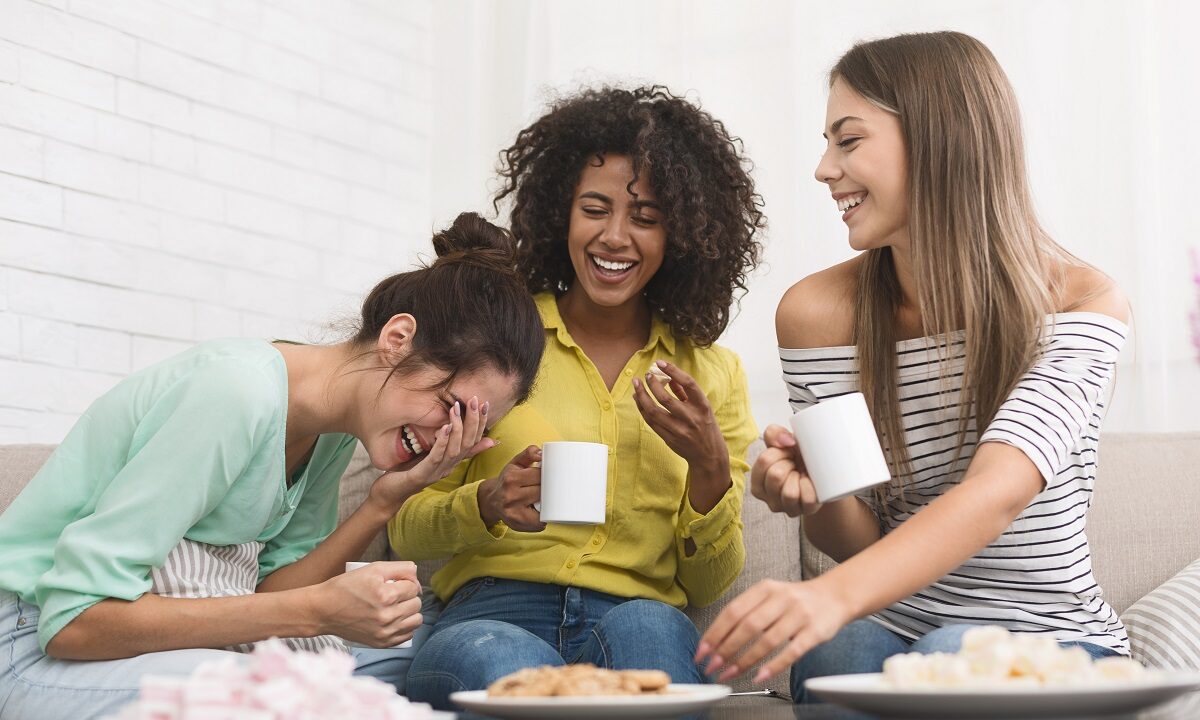Three young female roommates drinking coffee, eating cookies and laughing in their apartment.