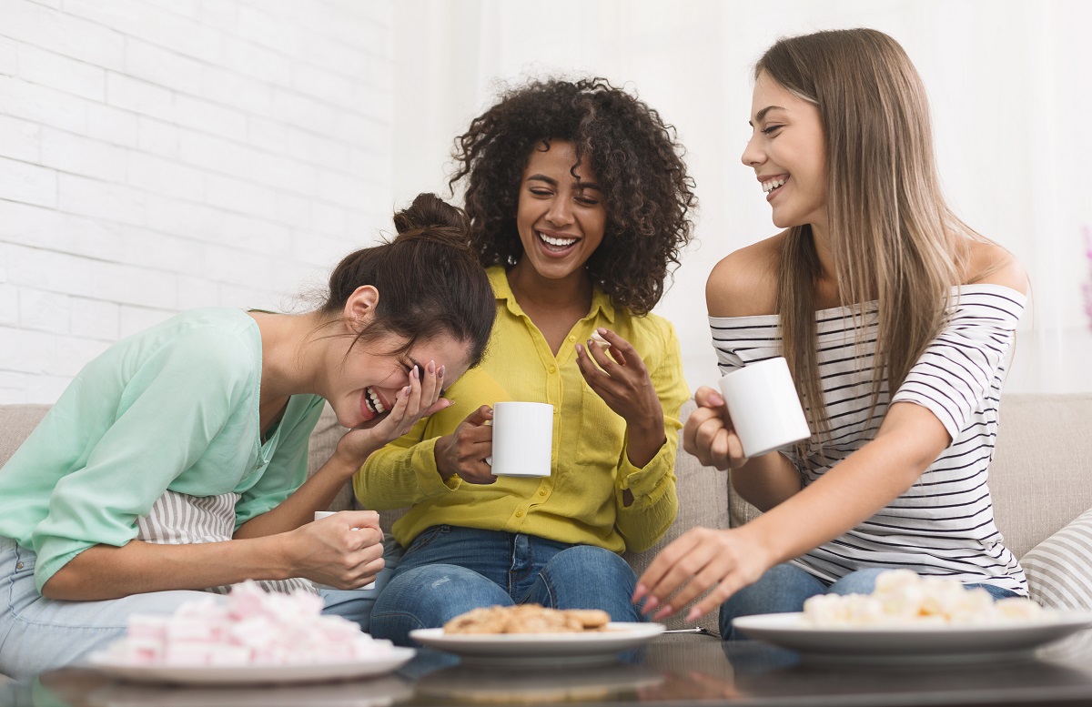 Three young female roommates drinking coffee, eating cookies and laughing in their apartment.