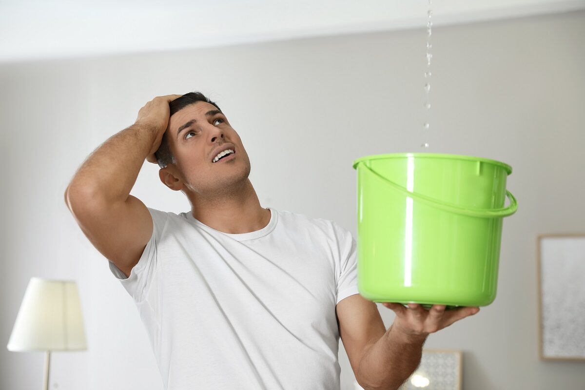 orried young man collects water in a green bucket from the leaking ceiling in the living room.