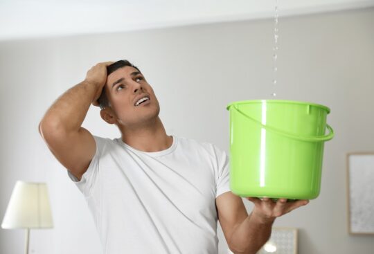 orried young man collects water in a green bucket from the leaking ceiling in the living room.
