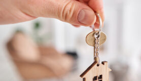 A man’s hand holds a key with a house-shaped keychain.