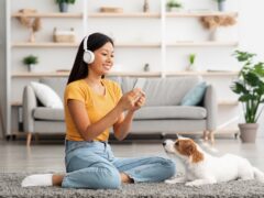 Cheerful young woman sitting on the floor in the living room with a mobile phone, taking a photo of her dog.