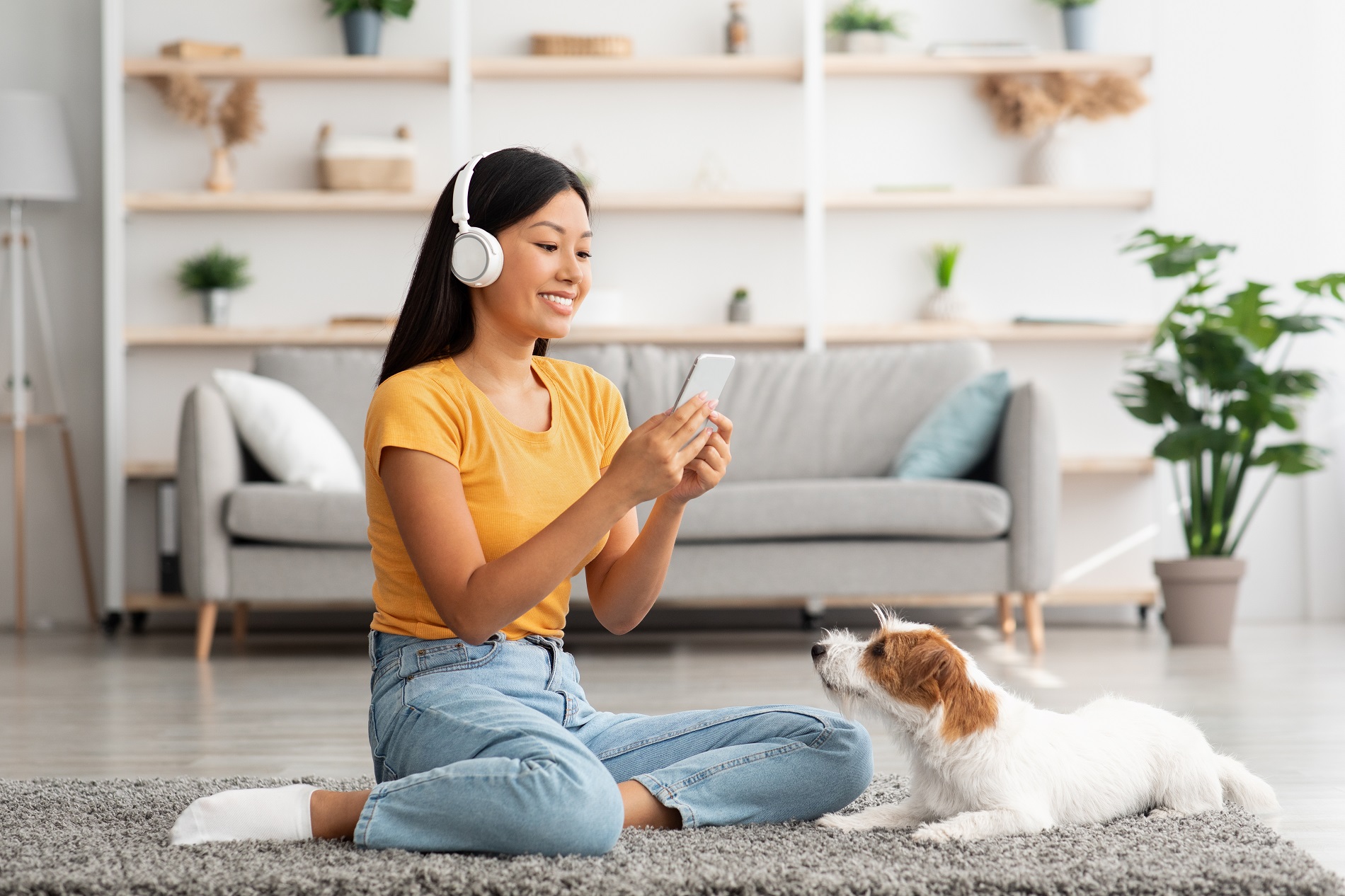 Cheerful young woman sitting on the floor in the living room with a mobile phone, taking a photo of her dog.