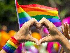 Supporting hands make heart sign and wave in front of a rainbow flag flying on the sidelines of a summer gay pride parade