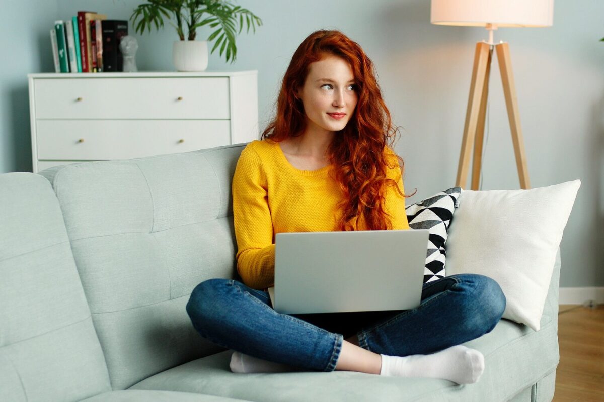 Cheerful redhead woman uses a silver laptop while sitting on a sofa in the living room in her apartment.