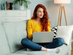 Cheerful redhead woman uses a silver laptop while sitting on a sofa in the living room in her apartment.
