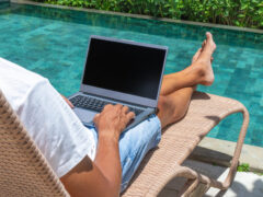 A man uses his laptop by a swimming pool during a workcation.