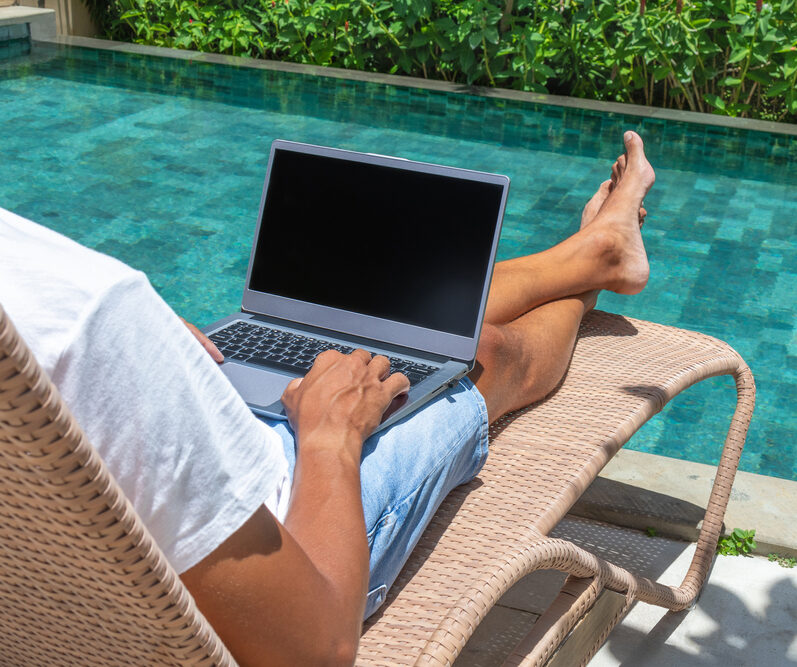 A man uses his laptop by a swimming pool during a workcation.