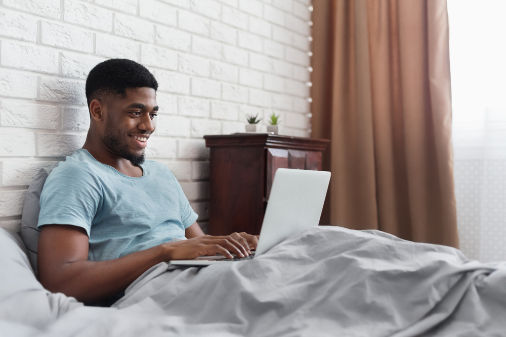 Always online. Black waking man working on laptop in bed, copy space. Early morning. Pleasant awakening.