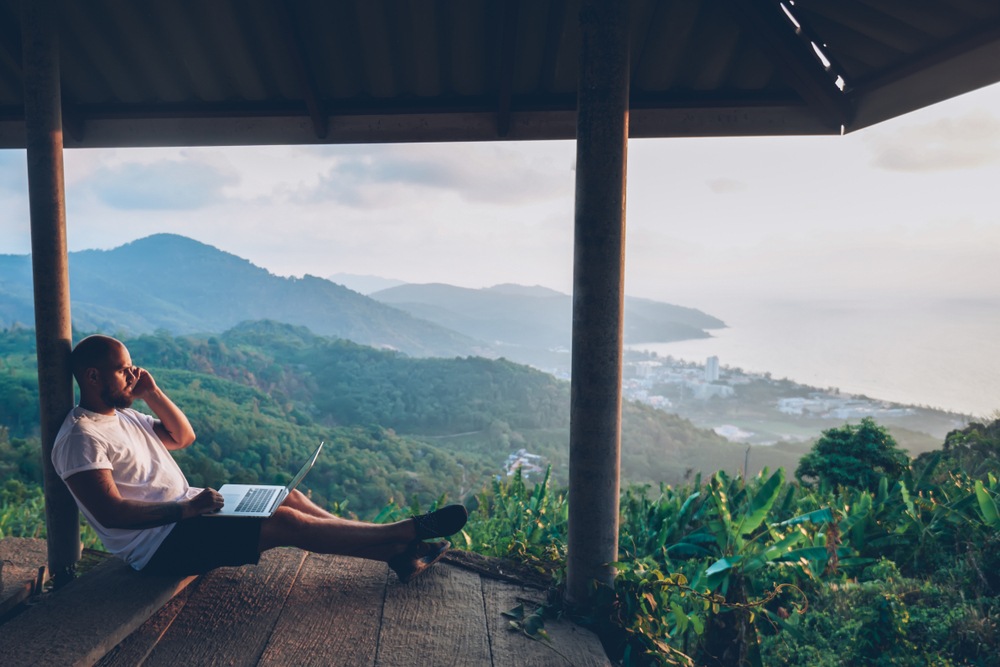 Young caucasian male freelancer talking on mobile phone while working on laptop computer remote. Hipster gut traveler working distantly while enjoying amazon nature landscape during summer vacations