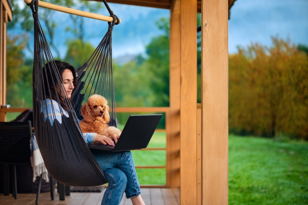 Young freelancer woman with dog working at the computer on the hanging chair on the terrace. Concept of the workplace at home, working remotely.