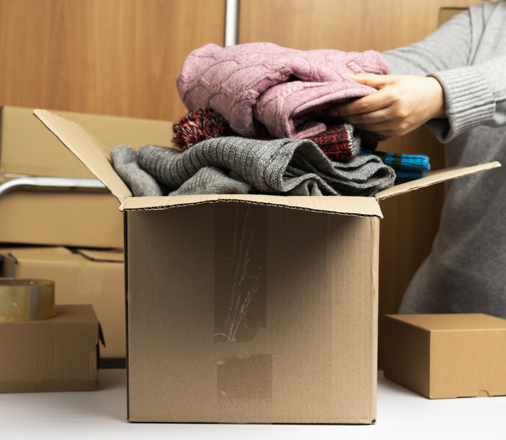 woman in a gray sweater collects clothes in a box, concept of assistance and volunteering, moving. Behind a stack of brown boxes