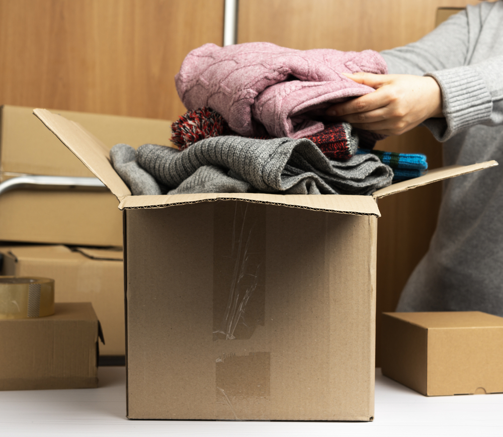 woman in a gray sweater collects clothes in a box, concept of assistance and volunteering, moving. Behind a stack of brown boxes woman in a gray sweater collects clothes in a box, concept of assistance and volunteering, moving. Behind a stack of brown boxes