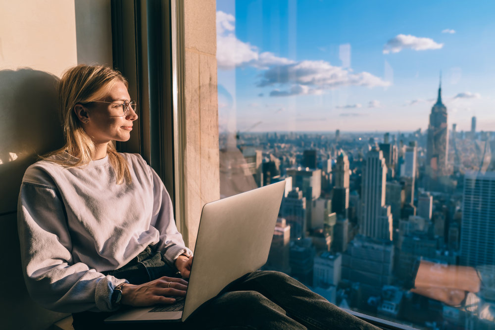 Caucasian female digital nomad sitting with laptop computer on window sill of modern skyscraper building coworking. Young woman freelancer enjoying downtown cityscape from hotel room during vacations