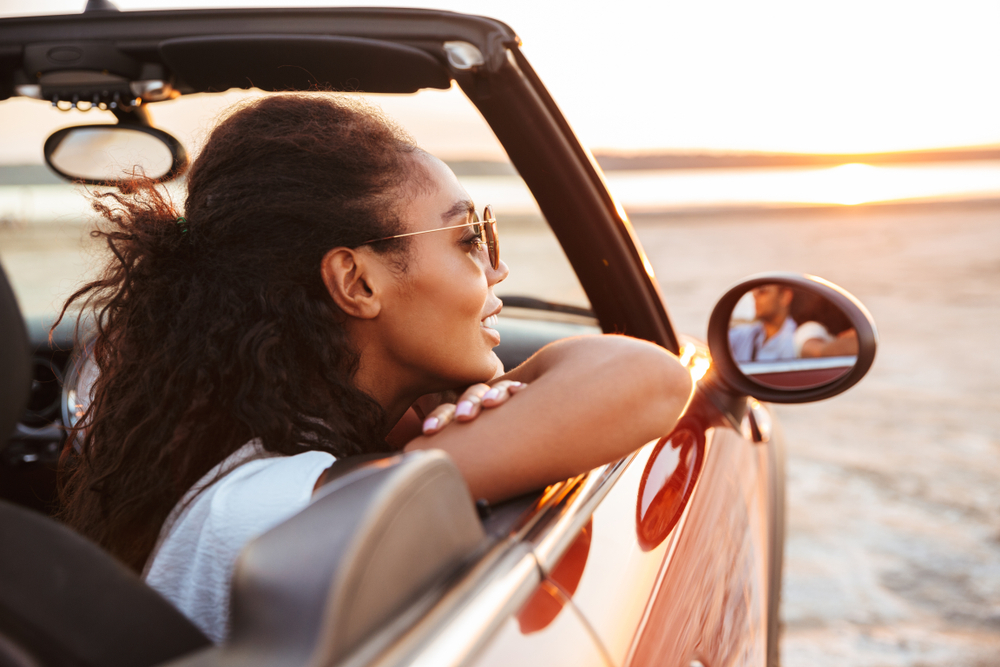 Image of young pretty woman smiling and looking aside while riding in convertible stylish car by seaside Image of young pretty woman smiling and looking aside while riding in convertible stylish car by seaside