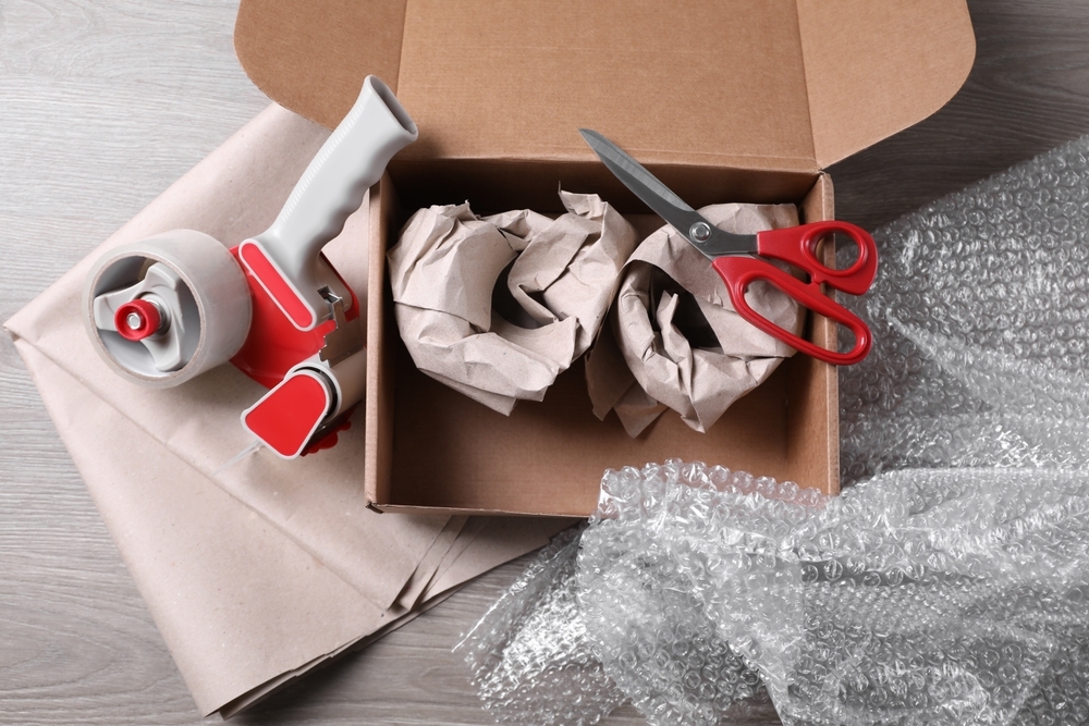 Open box with wrapped items, adhesive tape, scissors, paper and bubble wrap on wooden table, flat lay