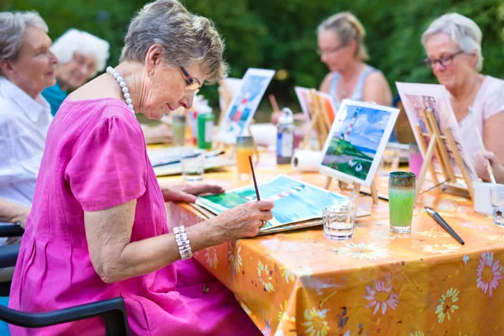 Stylish senior lady painting in art class with friends from her care home for the aged copying a painting with water colors on a canvas outdoors at a table in the garden. Stylish senior lady painting in art class with friends from her care home for the aged copying a painting with water colors on a canvas outdoors at a table in the garden.