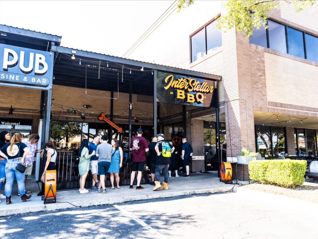 People in line at the front of a barbecue restaurant with the sign Interstellar BBQ.