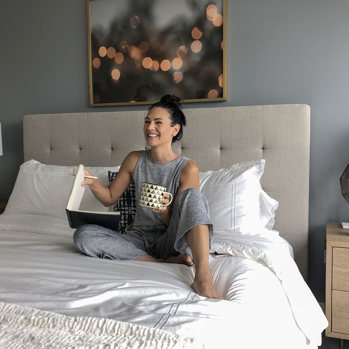 Woman with a notebook and a mug of coffee on a made bed in a furnished apartment